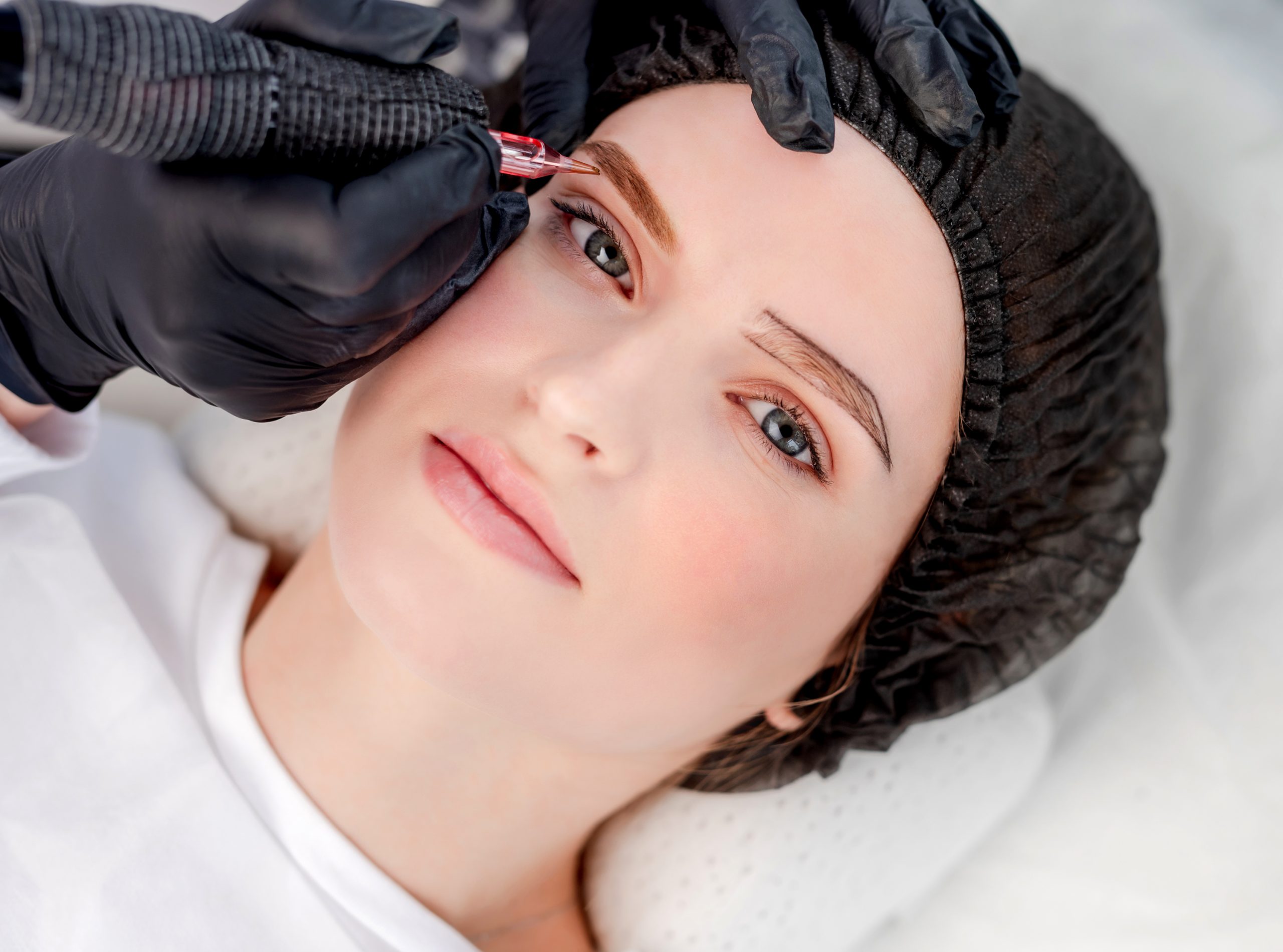 Beautiful model girl looking at camera during permanent eyebrows makeup process. Cosmetologist makes microblading
